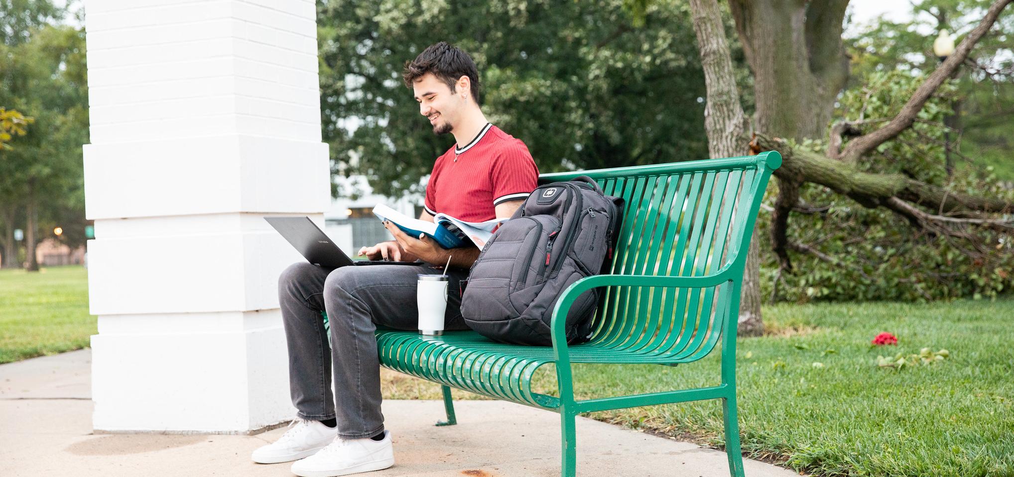 Student sitting on a green bench, holding book in hand while working on a laptop.