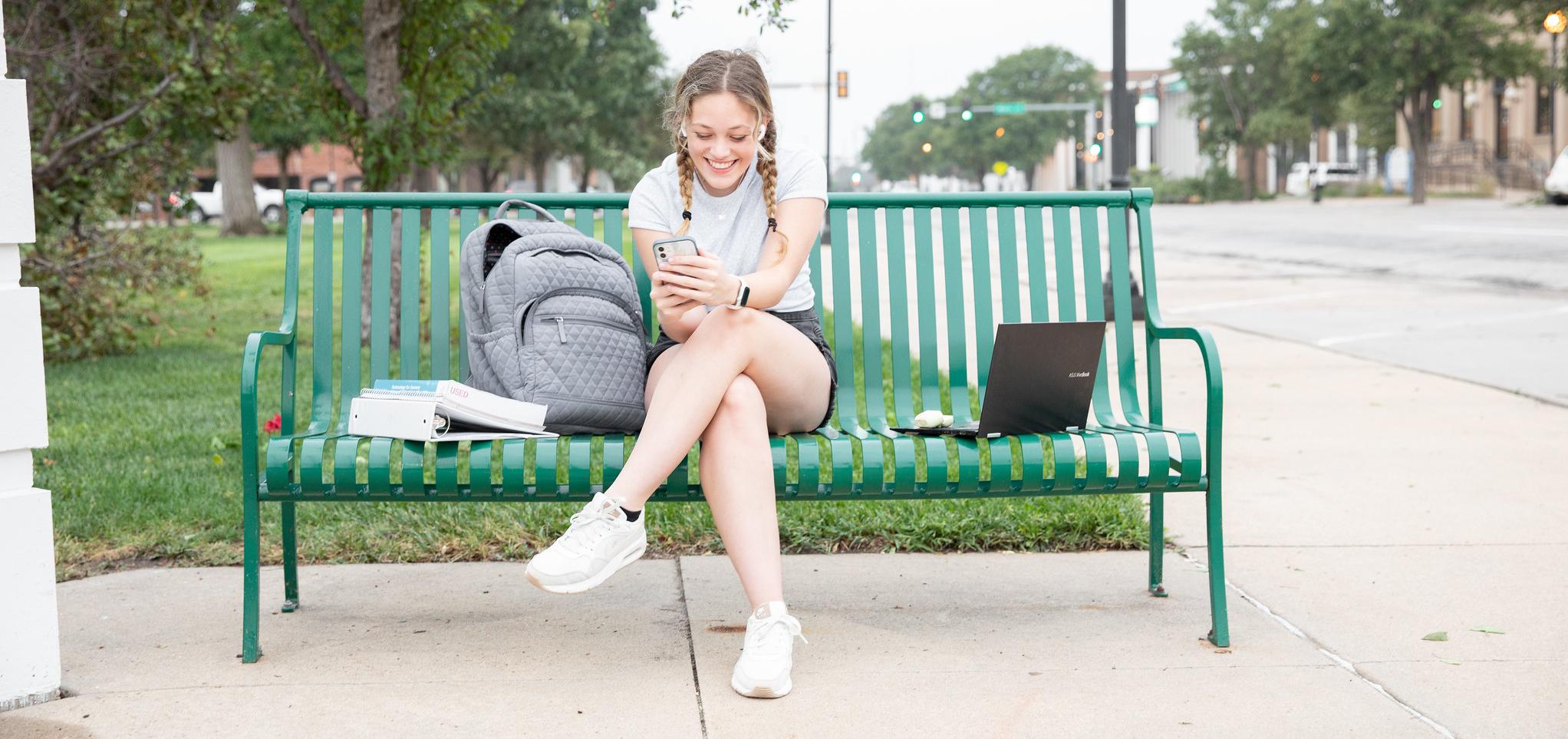 Student sitting on a green bench, with their phone in hand