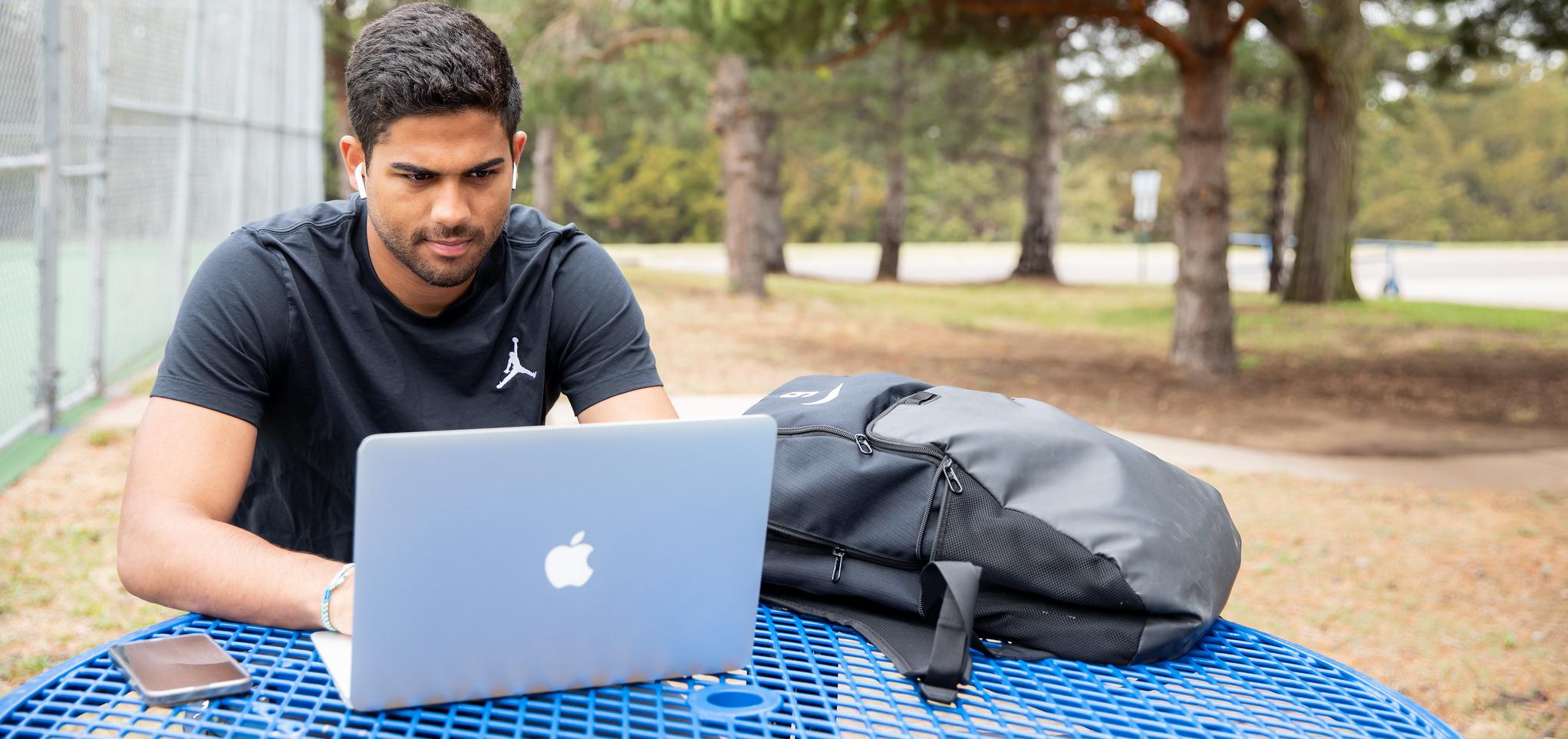 Student sitting in a park at a picnic table. There is a backpack, open laptop, and cell phone on the table.