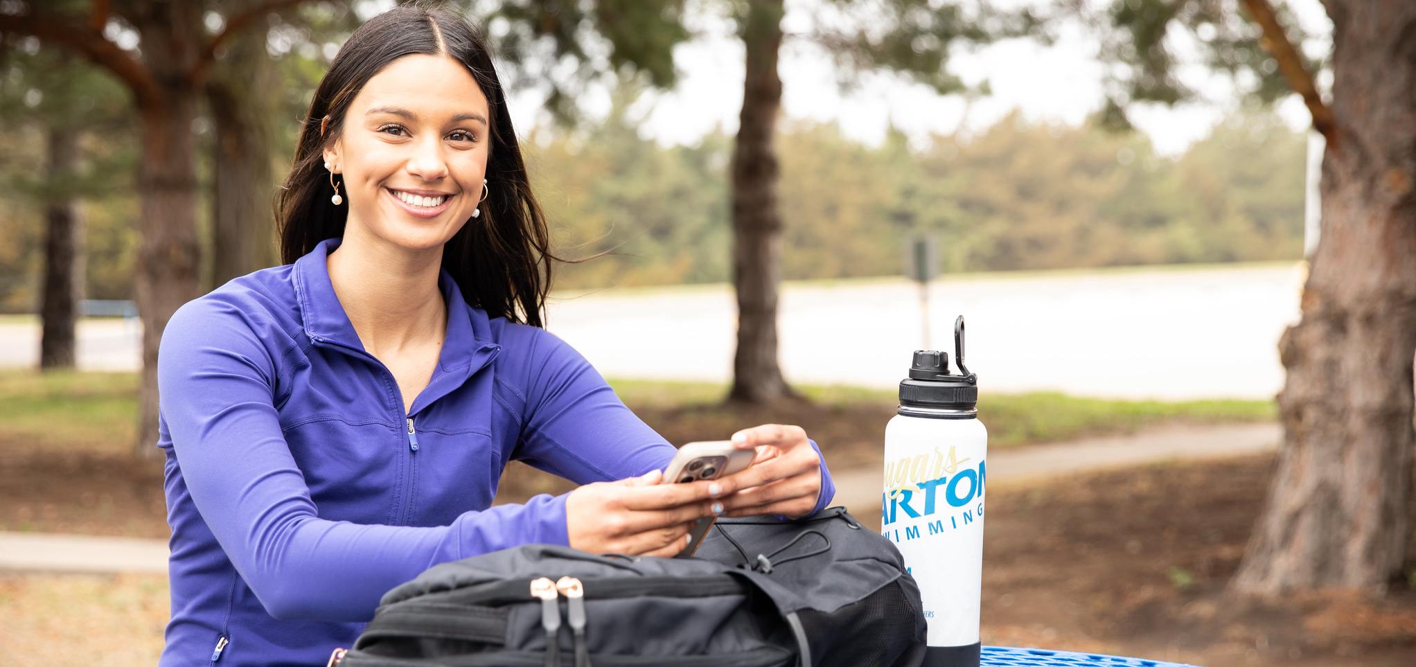 Female Student sitting at a picnic table with a backpack and water bottle on the table and phone in her hand. 