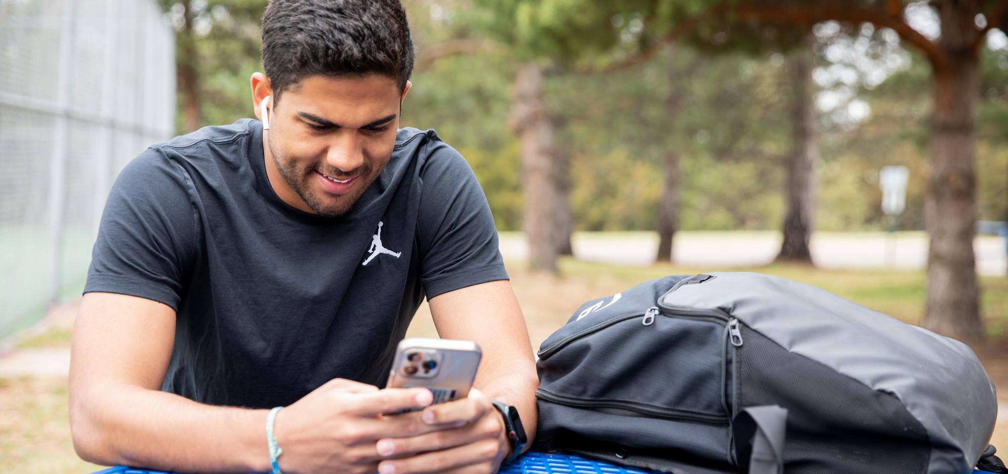 Student is sitting at a picnic table looking at a phone smiling with a backpack on the table