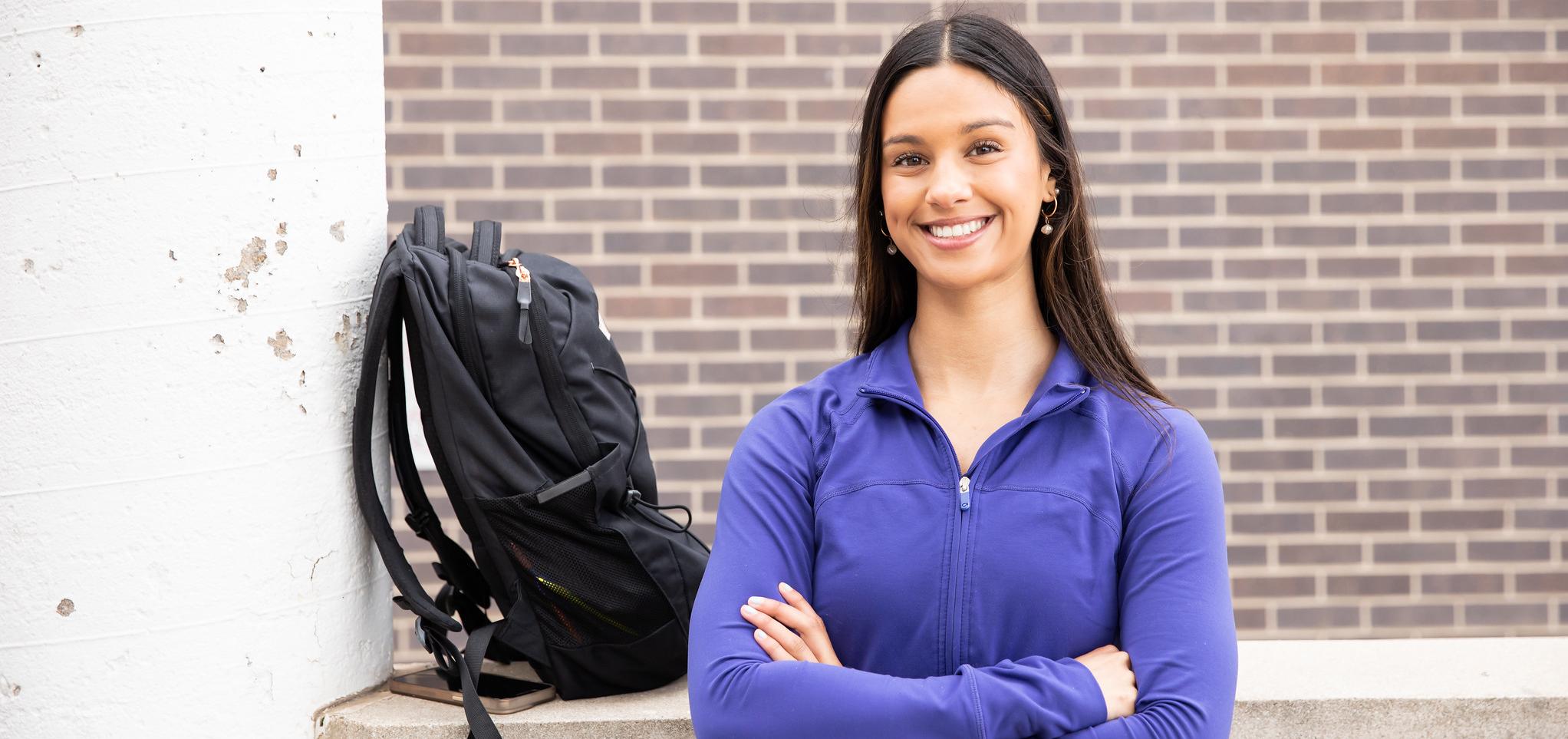 Student standing in front of wall with arms crossed and backpack on the wall.