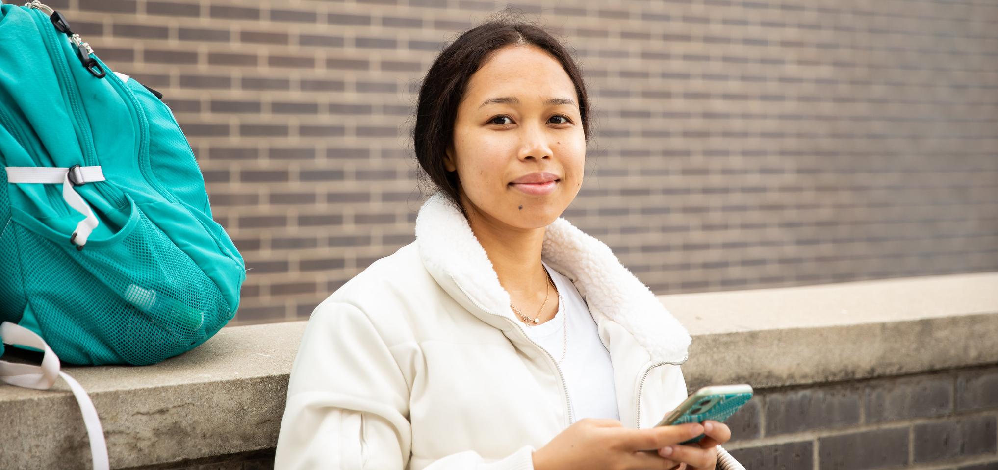 A student is holding their phone looking at the camera, while their backpack sits on a wall behind them. 