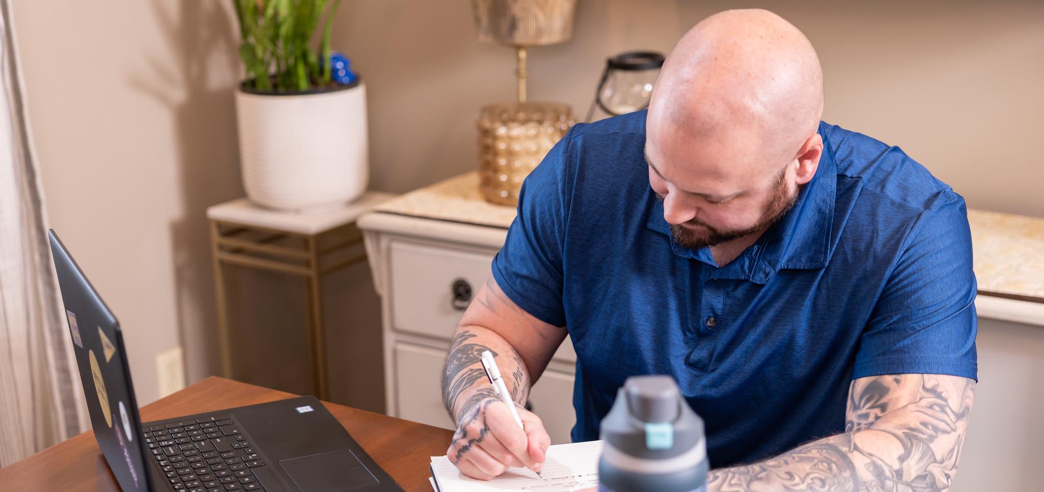 A bald man with tattoos writing in a notebook at a table with a laptop and water bottle.