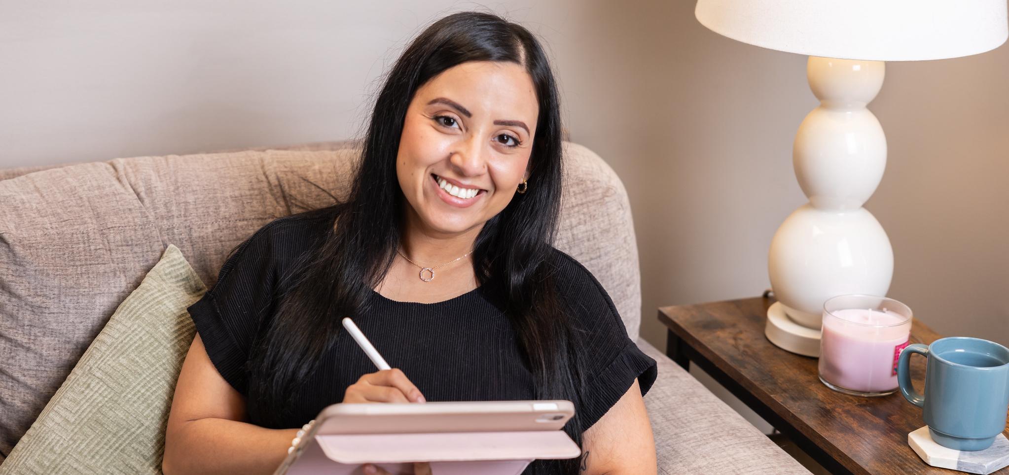 Woman smiling while using a tablet on a beige couch near a lamp and a side table.